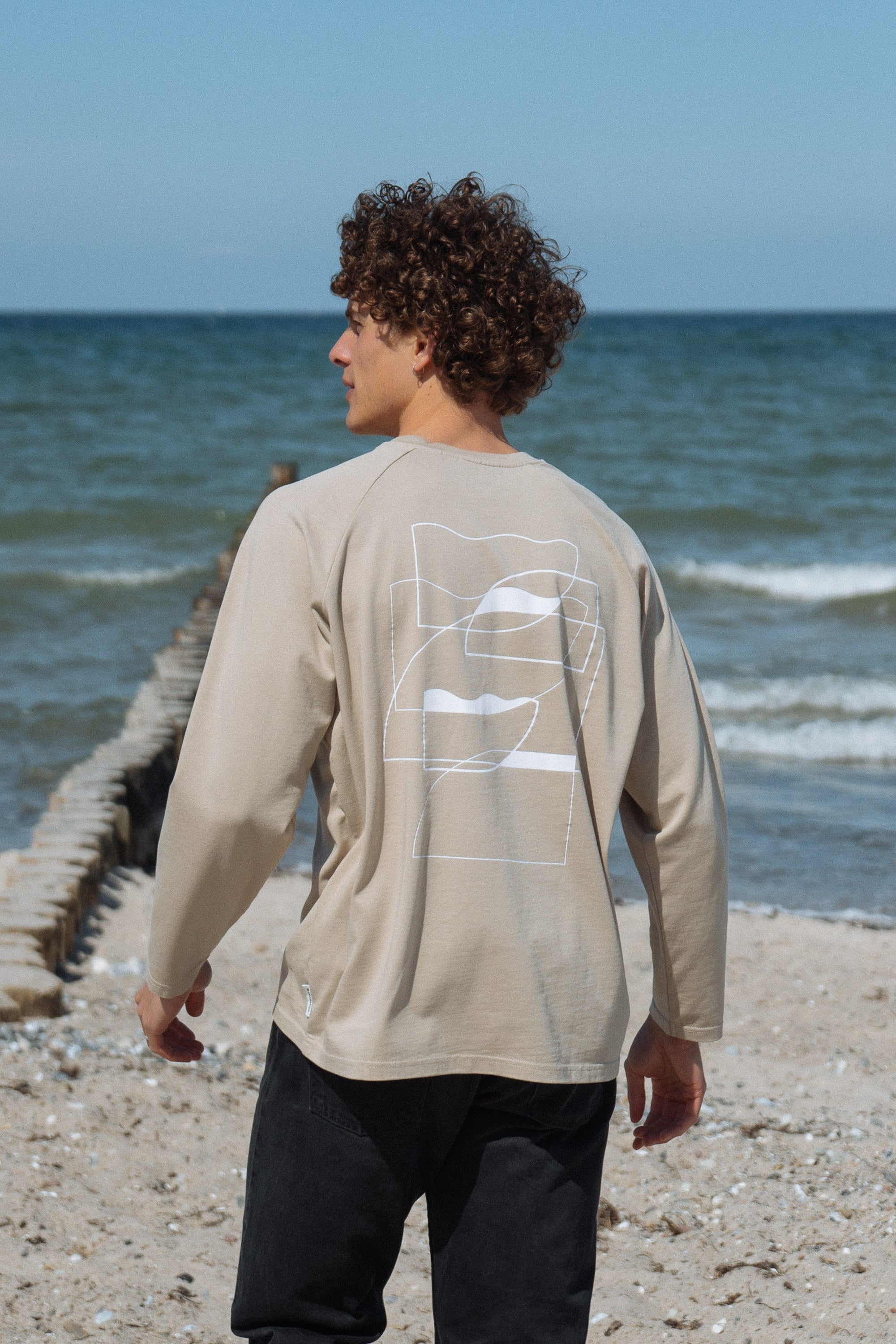 Mann trägt Statement Longsleeve in Sand von SALZWASSER am Meer _men